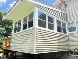 Exterior corner view of a sunroom addition with white-framed windows and tan vinyl siding, attached to the main house with a retracted awning above