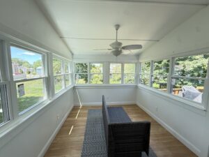 Vaulted sunroom interior with rows of white-framed double-hung windows on three walls, wood-look flooring, a ceiling fan, and dark wicker furniture