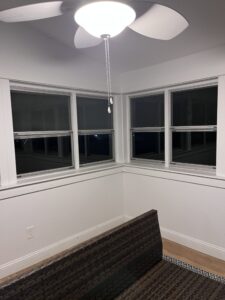 Corner of a room at dusk with two white-framed double-hung windows above wainscoting and wicker furniture in the foreground