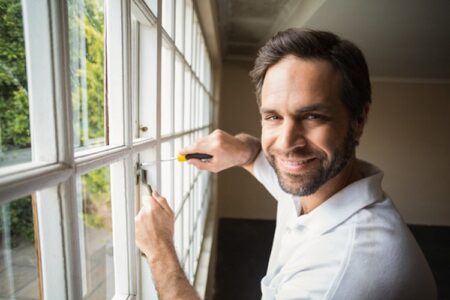 Smiling man in a white polo shirt using a screwdriver to adjust the hardware on a white double-hung window from inside a room
